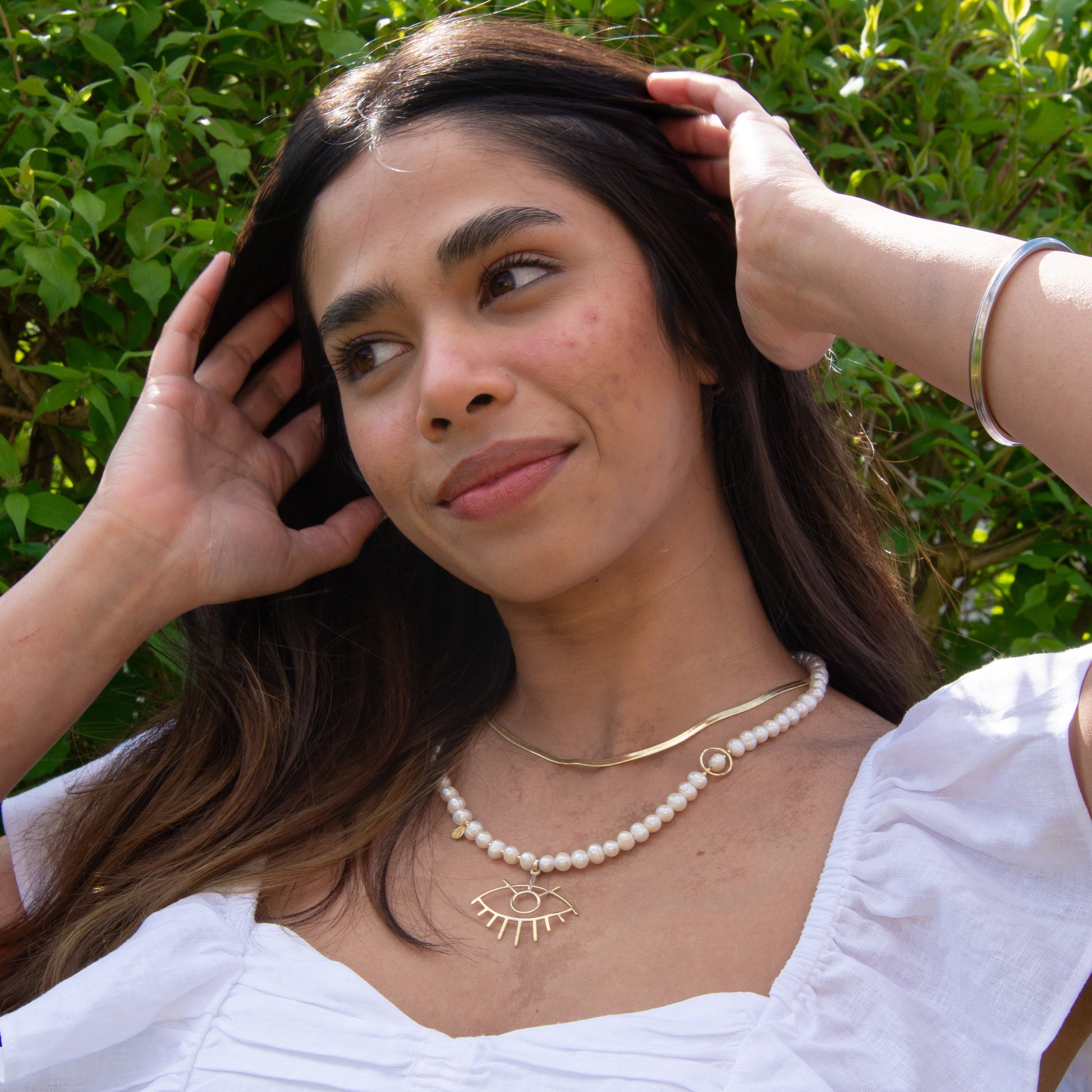 Woman wearing a Baroque Pearl Silver Eye Necklace with gold detail, looking relaxed in a garden setting.