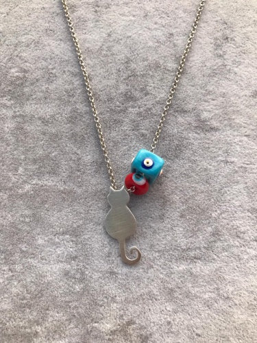 Silver necklace featuring a cat silhouette pendant alongside a blue and red Evil Eye charm on a gray background.