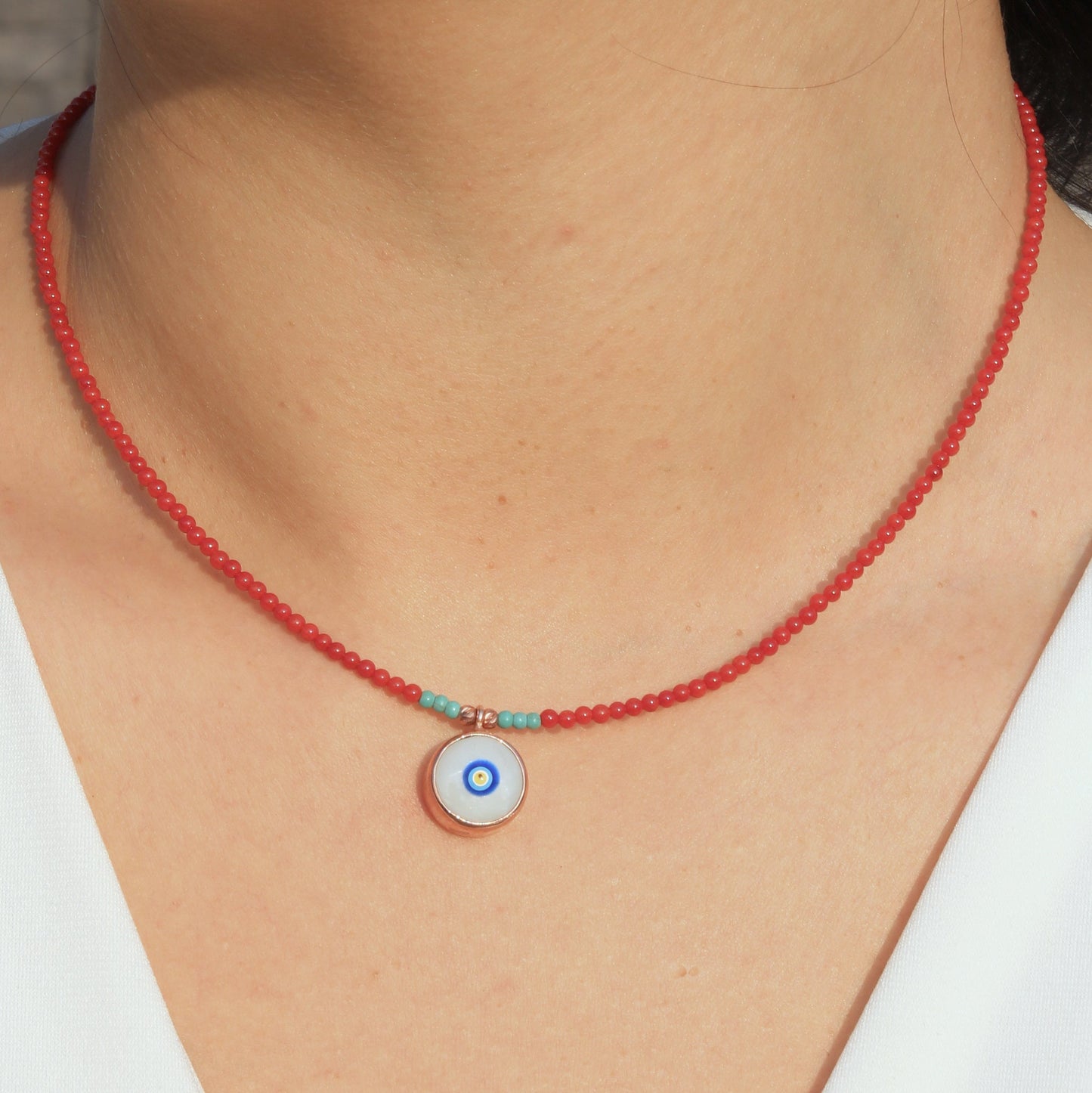 Close-up of a woman wearing an Evil Eye Coral Gemstone Necklace featuring a red beaded chain with a blue and white evil eye pendant.