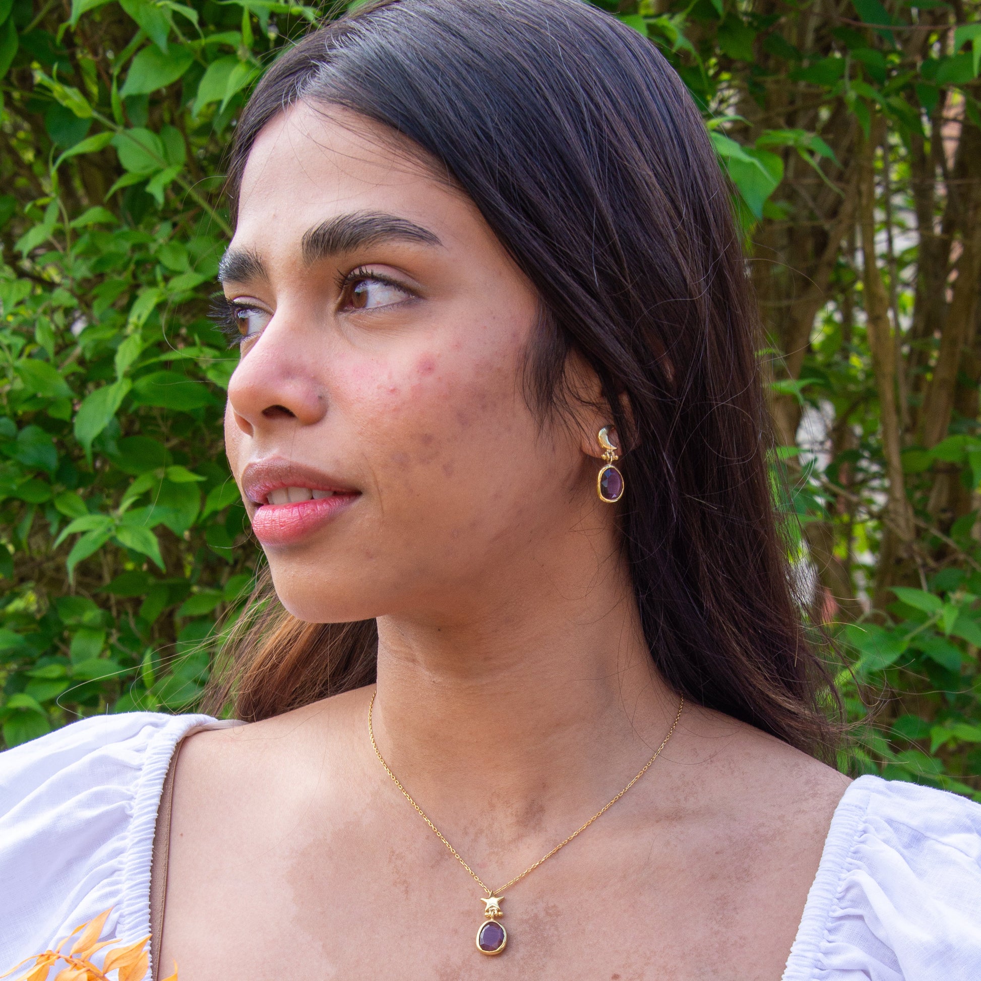 Woman wearing Foligno Purple Cat's Eye Gemstone Gold Plated Silver Chain Necklace and matching earrings, with a green foliage background.