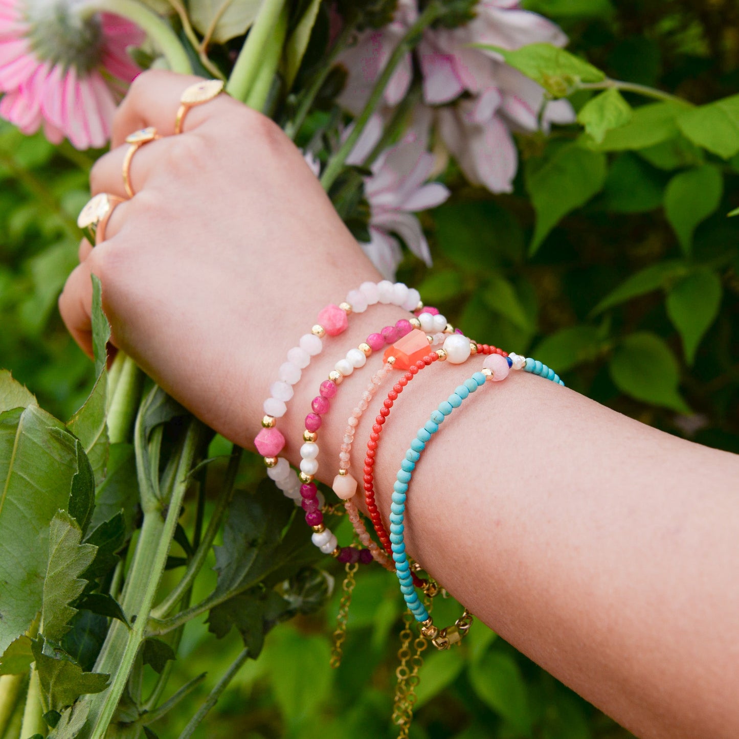 Colorful assortment of gemstone beaded and pearl bracelets on a wrist, holding pink flowers.