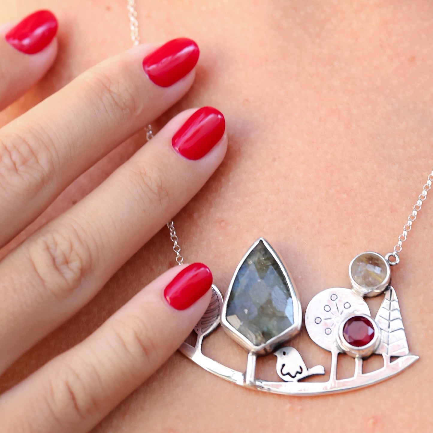 Close-up of a Landscape Silver Necklace with gemstones being worn, highlighted by red manicured nails.