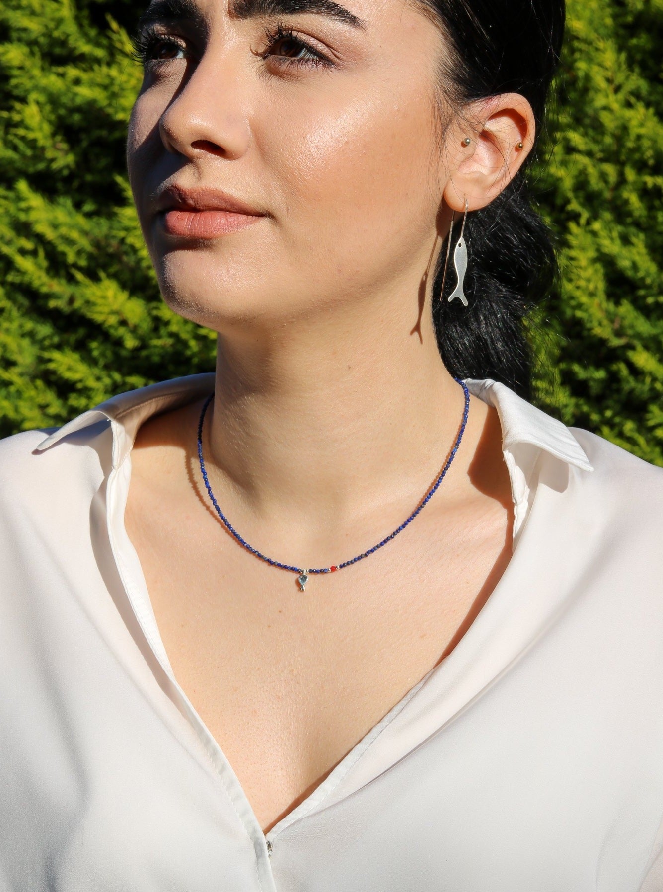 Woman wearing Minimal Lapis Lazuli Gemstone Silver Fish Necklace and matching silver fish earrings against a green foliage background.