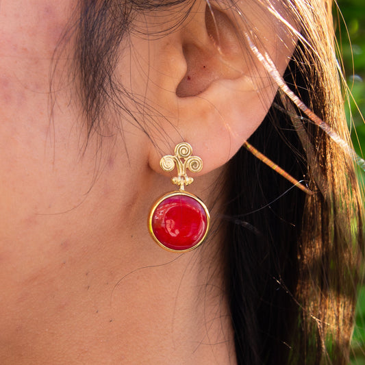 Close-up of a woman's ear adorned with Rho Natural Coral Gemstone Earrings featuring a round red coral stone set in gold.