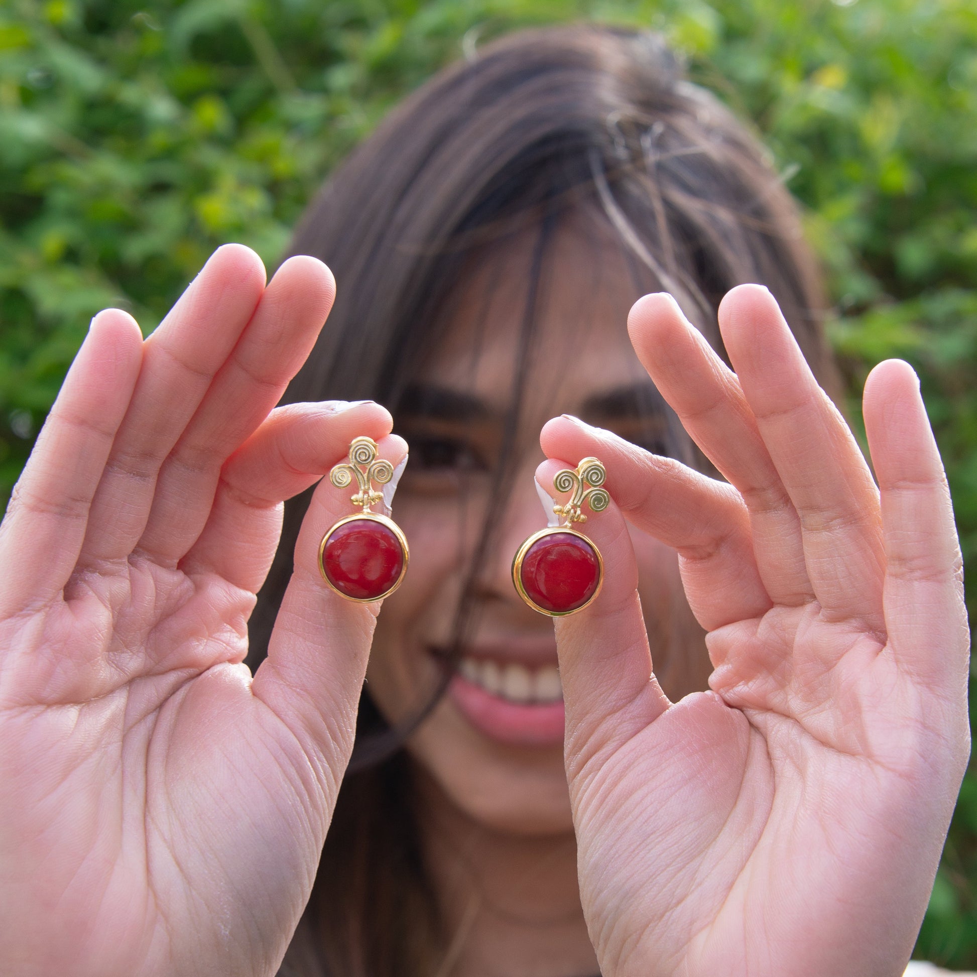 Woman holding Rho Natural Coral Gemstone Earrings against a green outdoor background.