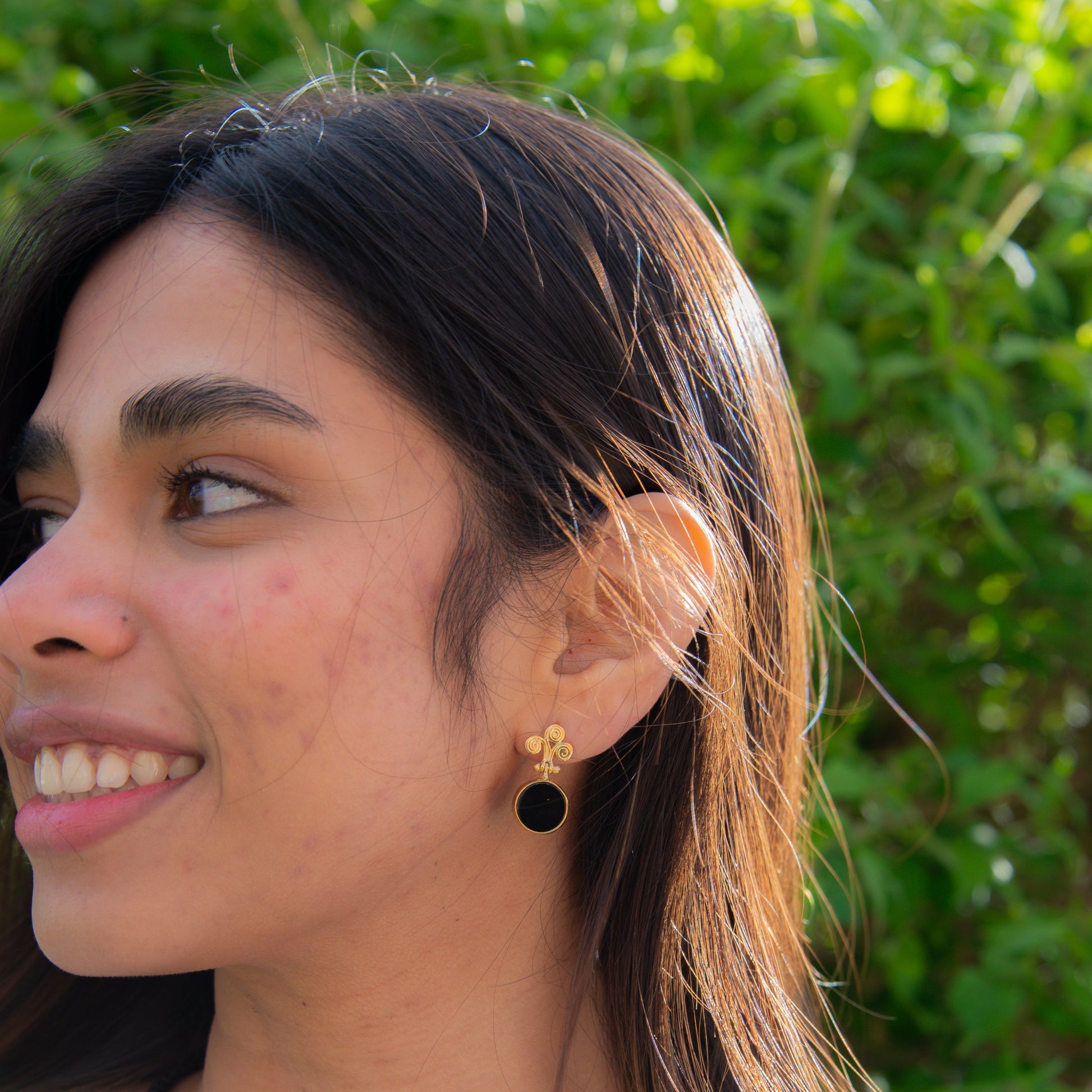 Woman wearing Rho Natural Onyx Gemstone Drop Earrings with a gold design, background of green foliage