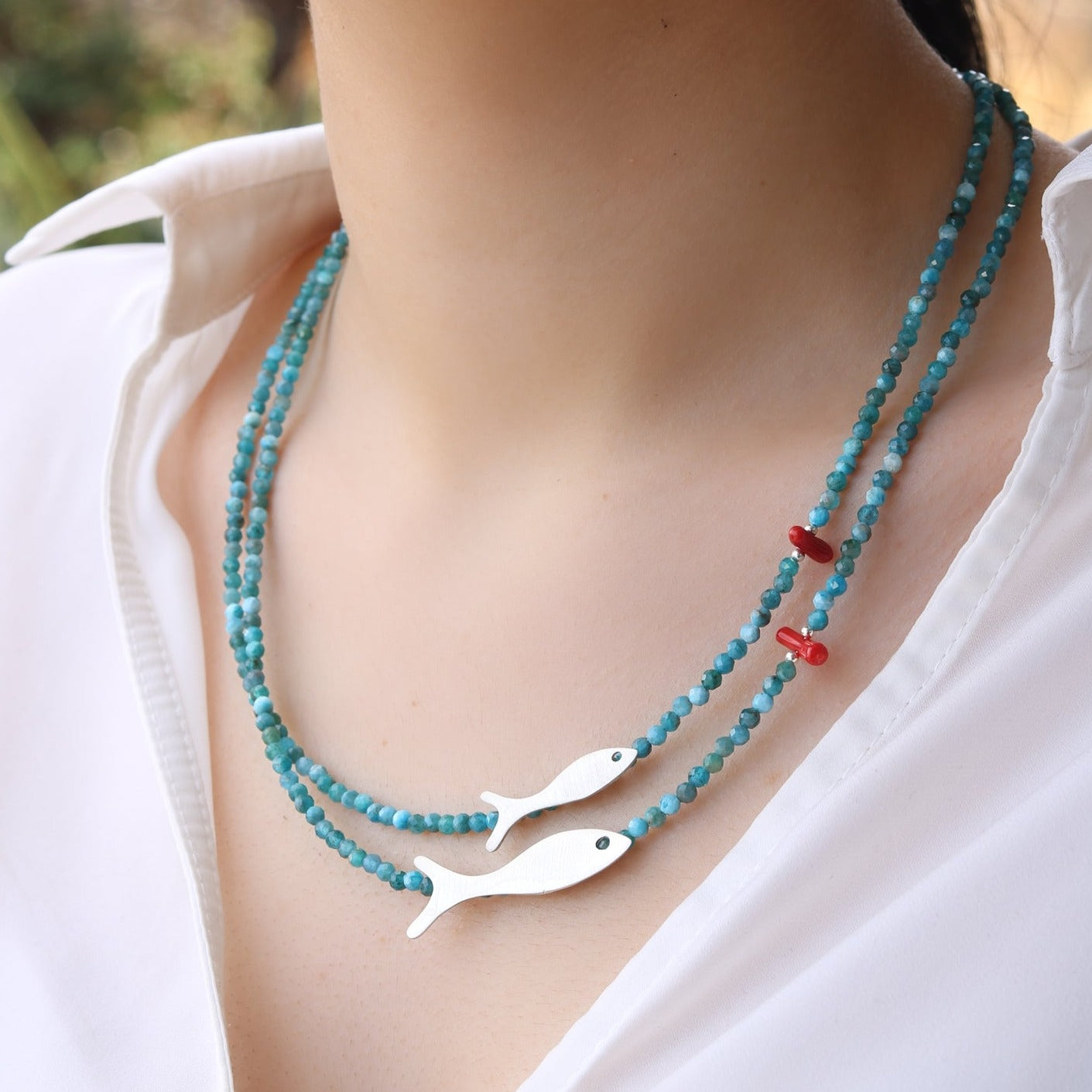 Close-up of Small and Big Fish Apatite Silver Necklace worn around the neck, featuring blue and red beads with silver fish pendants