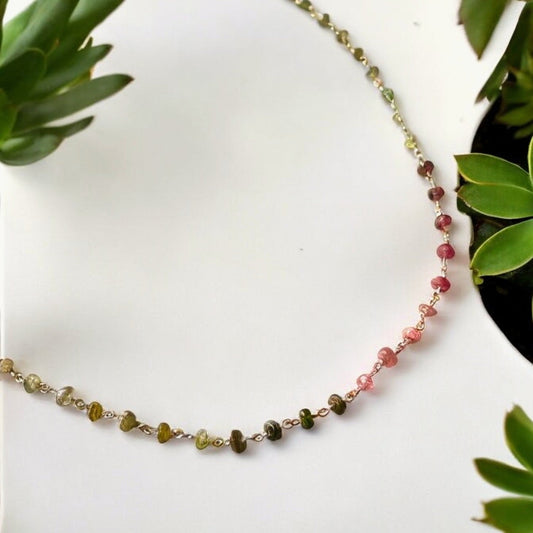 Tourmaline gemstone and silver necklace with mixed-color beads on white background surrounded by green plants